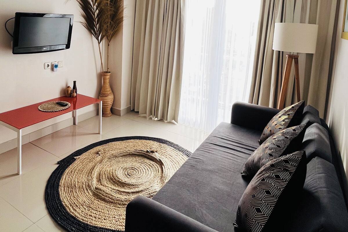 A modern living room with a sofa, a flat-screen TV mounted on the wall, a small table with a remote control, a floor lamp, and a large window with curtains.