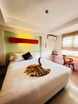 A hotel room with a neatly made bed, a desk with a chair, and a window with blinds. - Casa Kuta View 2 BR (Kuta)