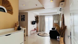 A modern living room with a white sofa, a coffee table, and a TV mounted on the wall. There is a large window with curtains, a decorative wall piece, and a white cabinet with drawers. - Casa Kuta View 2 BR (Kuta)