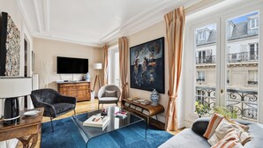 A modern living room with a sofa, coffee table, and armchairs. There is a large painting on the wall, a flat-screen TV, and a window with curtains.