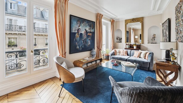A spacious living room with a large window, a blue carpet, a grey sofa, and a painting on the wall.