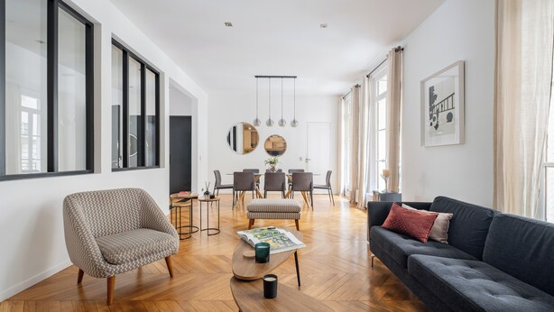 Un salon spacieux avec un canapé foncé, un fauteuil à motifs et une table basse en bois. On aperçoit à l’arrière-plan un coin repas avec une table ronde et des chaises.