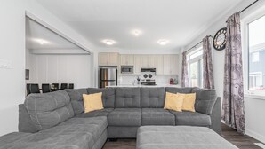Ein modernes Wohnzimmer mit einem grauen Ecksofa, einem Essbereich mit Tisch und Stühlen sowie einer Küche mit weißen Schränken und Edelstahlgeräten.
