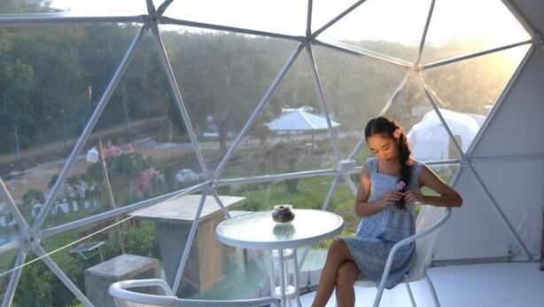 Une personne assise dans une pièce en forme de dôme géodésique, avec une table en verre et une vue sur un paysage.