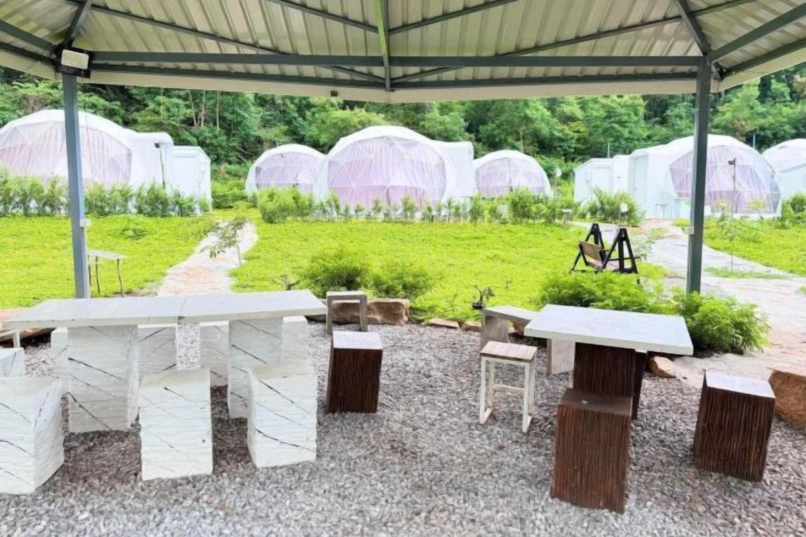 An outdoor covered seating area with tables and chairs, surrounded by a garden with white structures in the background.