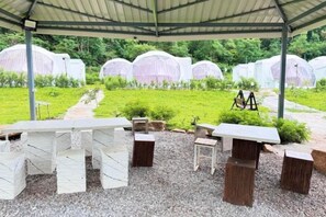 Un espace extérieur abrité, aménagé avec des tables et des chaises, entouré d’un jardin où se dressent, en arrière-plan, des structures blanches.