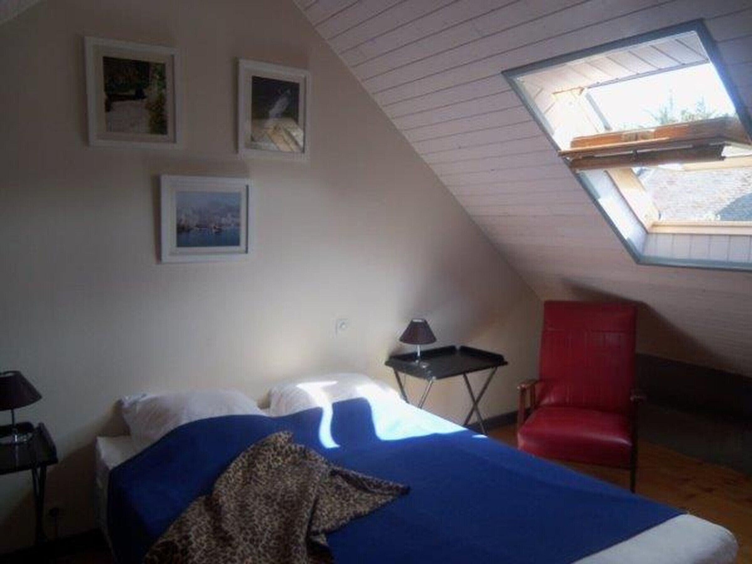 A bedroom with a sloped ceiling, a bed with a blue blanket, a red chair, and framed pictures on the wall.