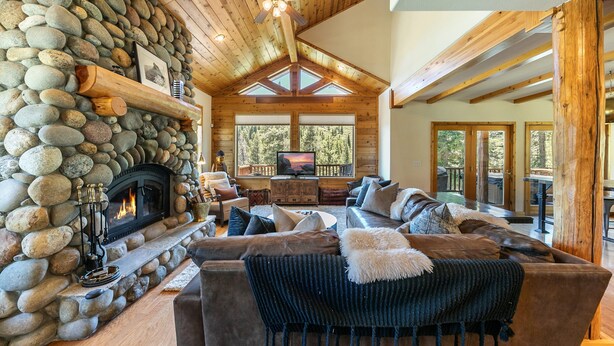 A cozy living room with a stone fireplace, wooden ceiling beams, and a large window overlooking a deck. - Horizon by Tahoe Getaways - Pet Friendly Tahoe Donner w Hot Tub (Truckee)