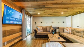 A modern living room with wooden paneling, a large flat-screen TV, a sofa, a coffee table, and a dining area. - Cozy chalet with Pool and Spa (Grenville-sur-la-Rouge)
