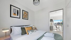 A hotel room with a bed, bedside tables, a lamp, artwork on the wall, and a view through an open door.