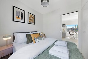 A hotel room with a bed, bedside tables, a lamp, artwork on the wall, and a view through an open door.
