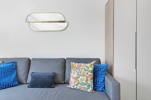 A grey sofa with three pillows, one floral and two blue, against a plain wall with a white door and a light switch.