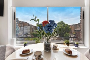 A dining table with a vase of flowers, plates with pastries, and a cup, set against a window overlooking a cityscape.