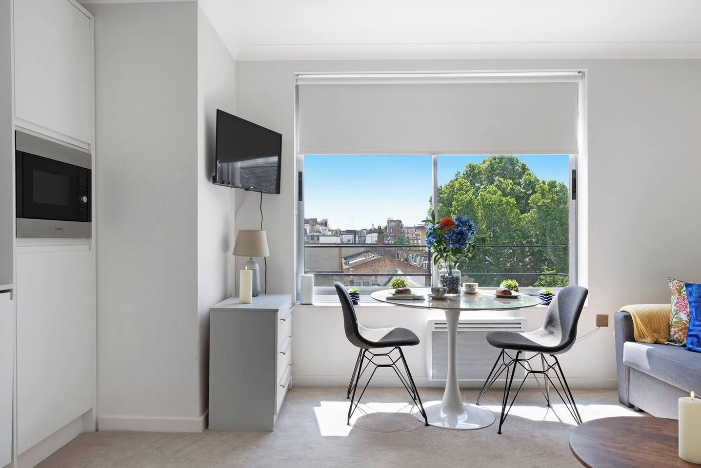 A modern apartment with a dining area, a flat-screen TV, a microwave, and a large window offering a city view.