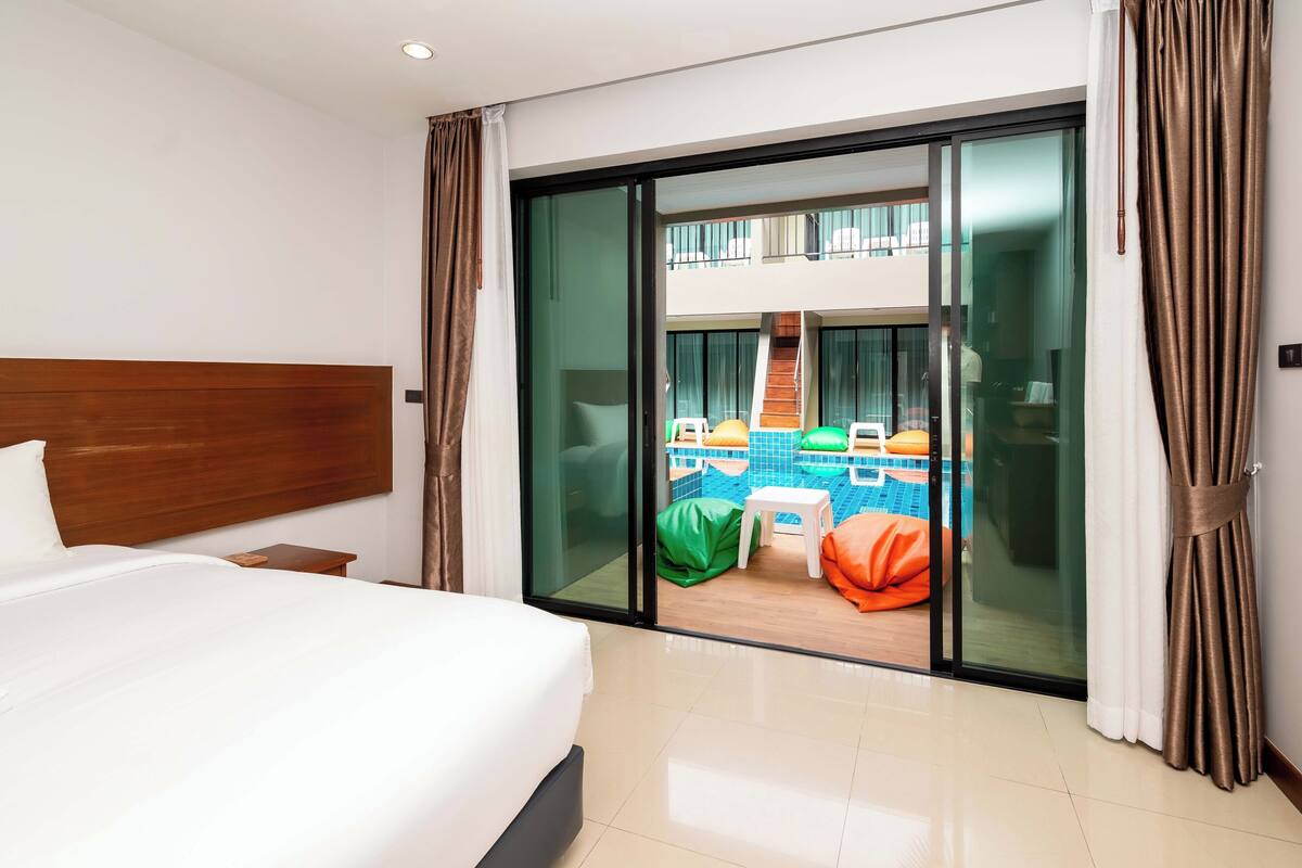 A hotel room with a bed, a nightstand, a sliding glass door leading to a pool area with colorful chairs and a small table.