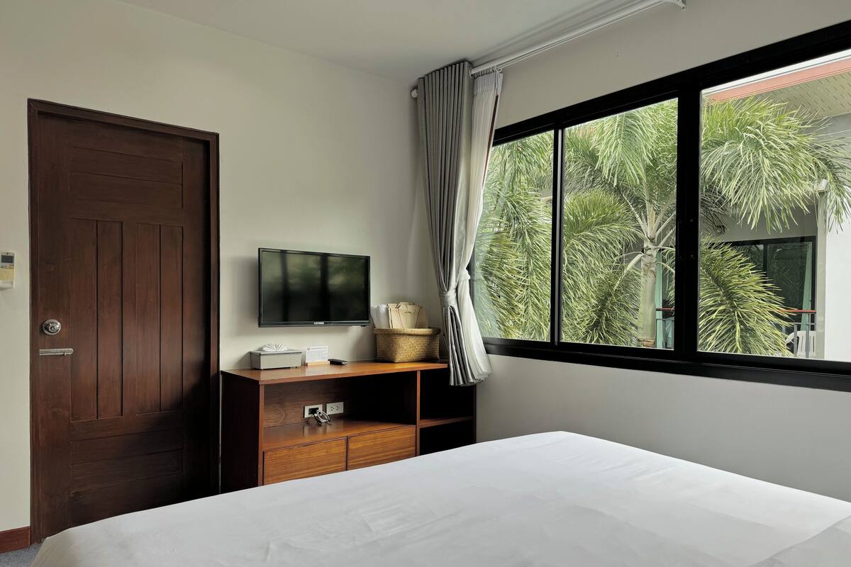 A hotel room with a bed, a TV on a wooden stand, a window with curtains, and a view of palm trees.