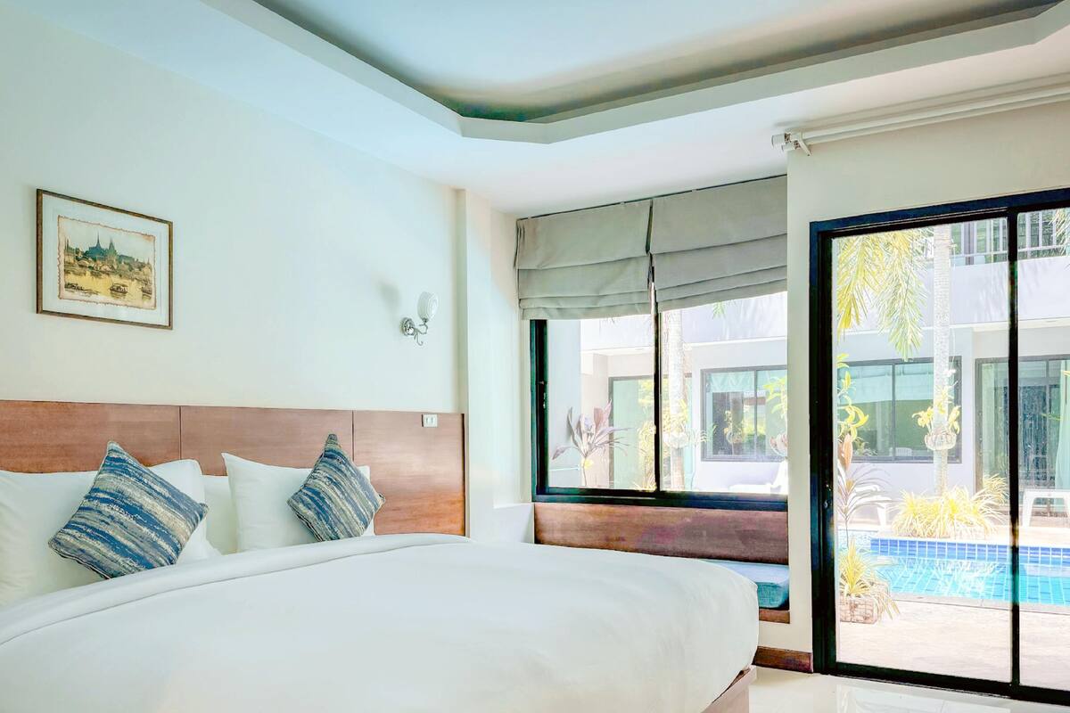 A hotel room with a large bed, a headboard, a window with a view of a pool and greenery, and a framed picture on the wall.