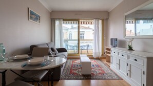 A modern living room with a sofa, dining table, and a TV. There is a balcony with a view of buildings.