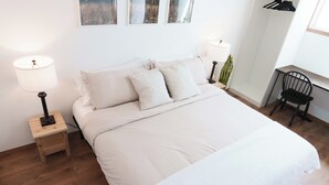 Un lit bien fait, avec du linge de lit blanc, une petite table de chevet en bois sur laquelle se trouve une lampe, et une chaise placée contre le mur.
