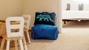 Une chambre à coucher avec un lit, une chaise et une petite table. Sur le lit, on trouve un oreiller orné d’un motif de dinosaure.