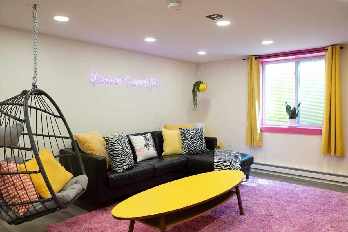A living room with a black sofa, yellow cushions, a hanging chair, and a neon sign on the wall.