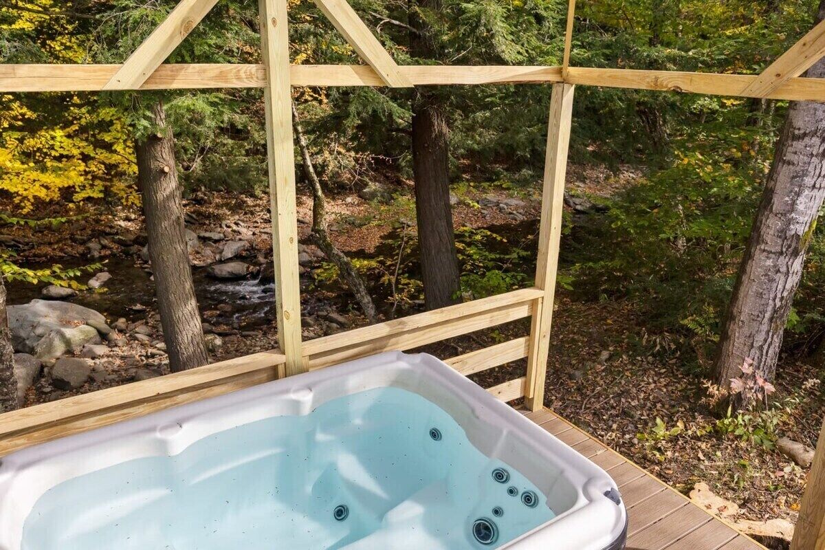 Un bain à remous installé sur une terrasse en bois, avec en arrière-plan un paysage naturel composé d’arbres et d’un ruisseau rocheux.