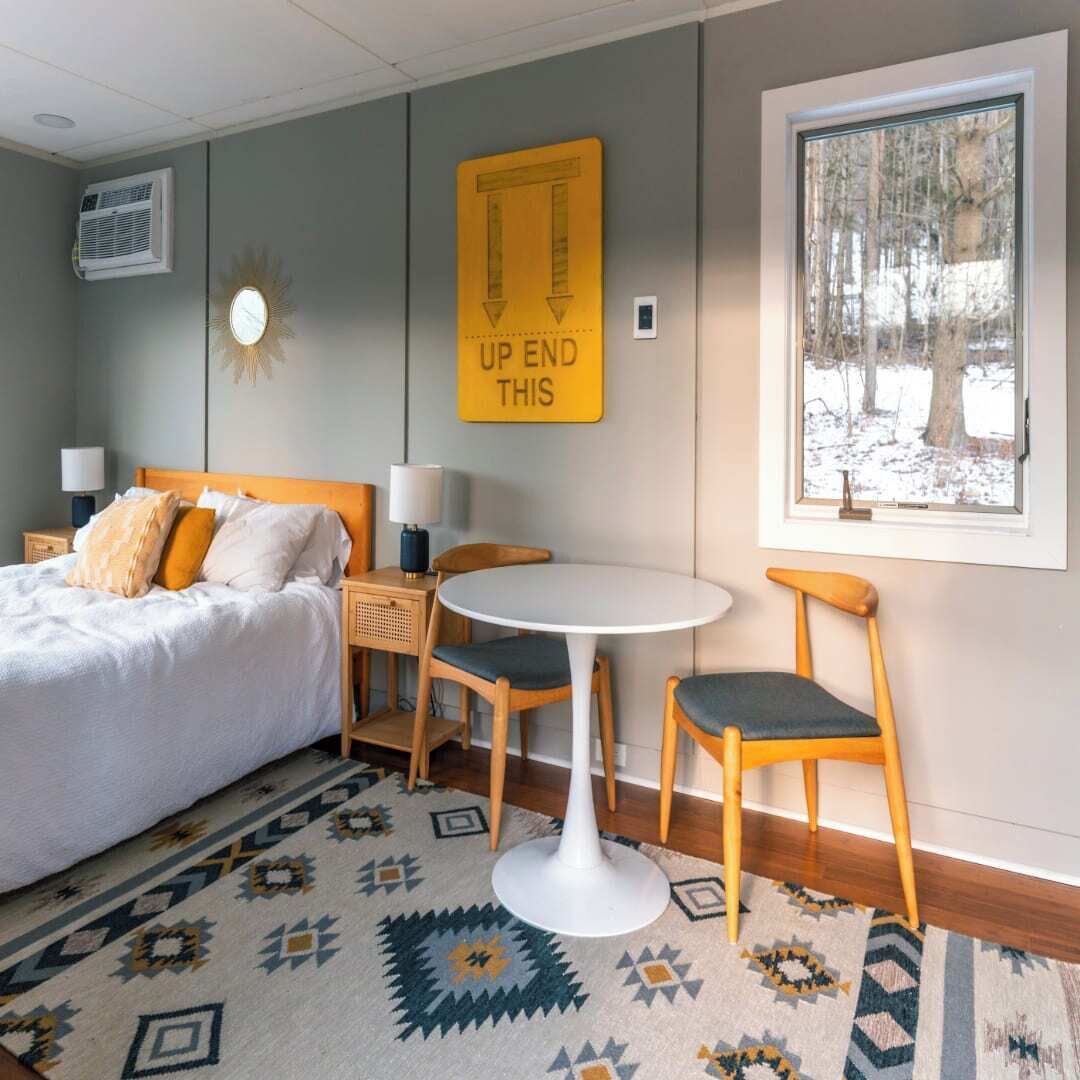 Habitación de hotel con cama, dos sillas, una mesita, ventana y vistas a árboles, además de un cartel en la pared.