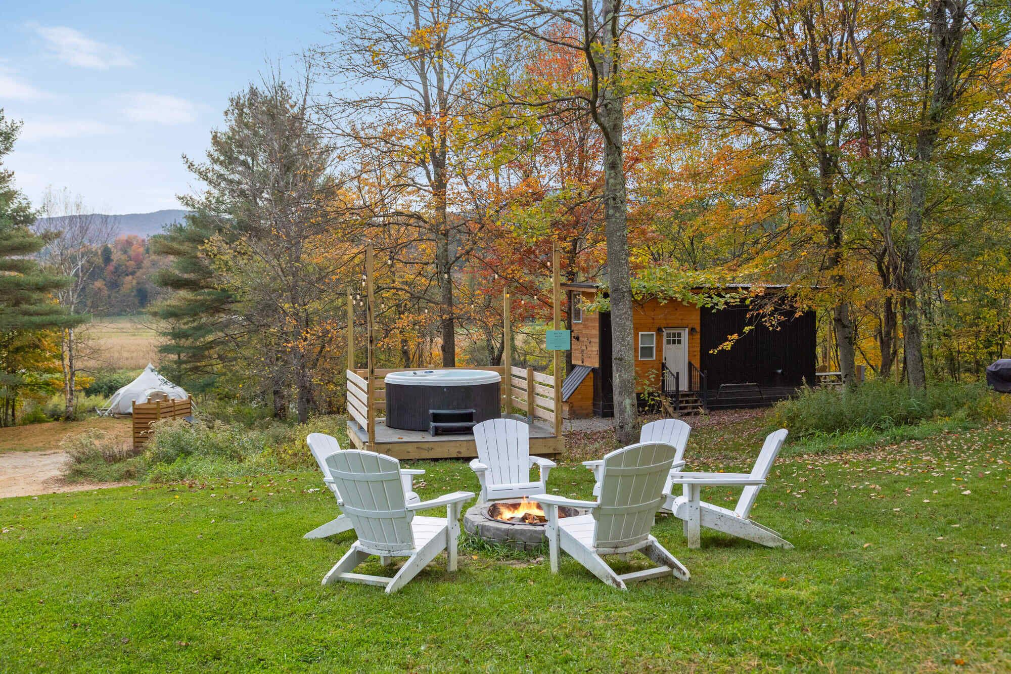 Ein gemütlicher Sitzbereich im Freien mit Adirondack-Stühlen um eine Feuerstelle, vor dem Hintergrund von Herbstlaub und einer rustikalen Holzhütte.