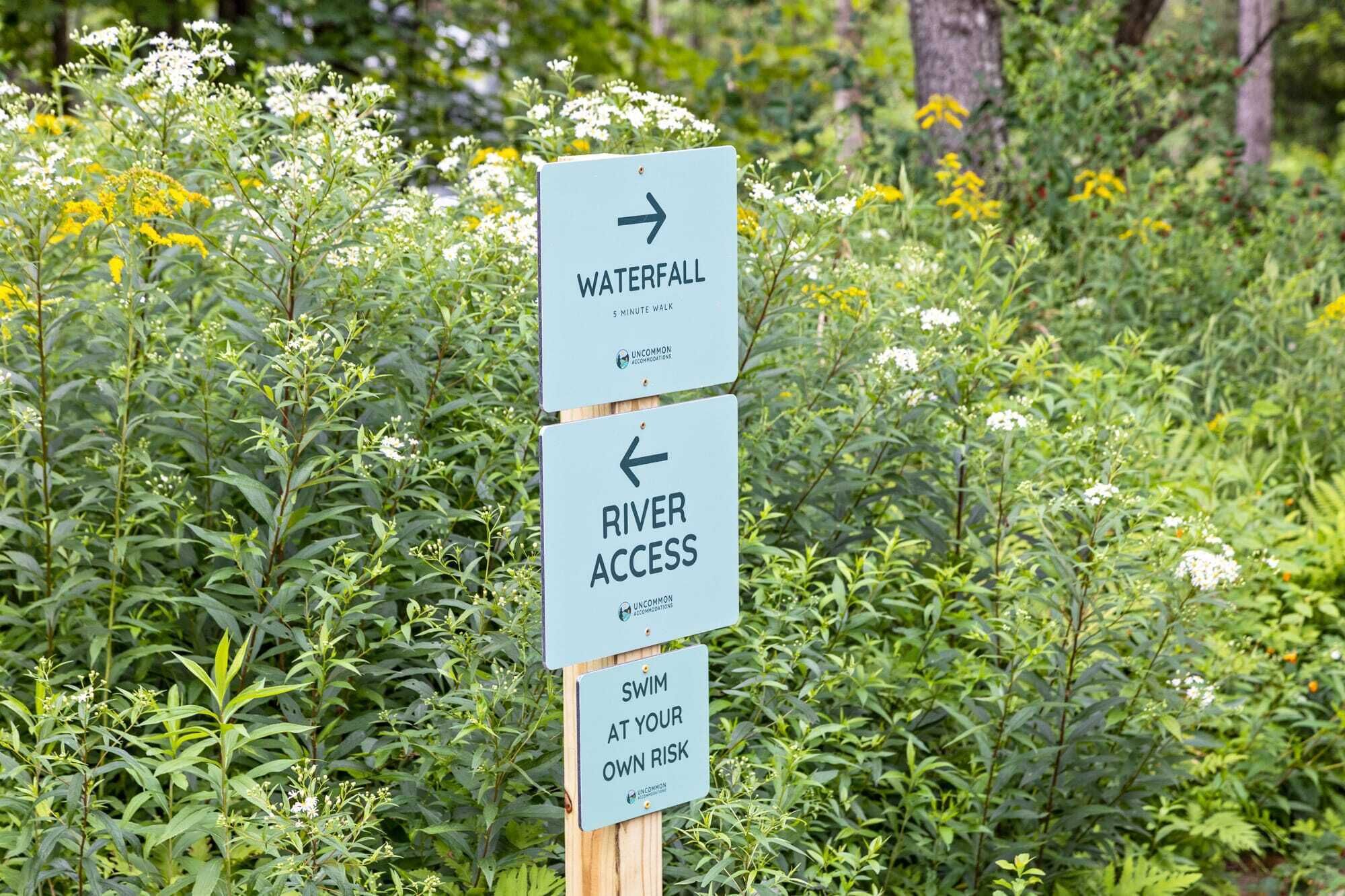 Un panneau indicateur indiquant la direction d’une cascade et d’un point d’accès à la rivière, ainsi qu’un avertissement indiquant que la baignade se fait à vos propres risques.