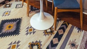 A hotel room with a round white table, two yellow mugs, a bed with white bedding, a patterned rug, and a yellow sign on the wall.