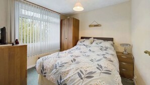 A bedroom with a bed, a bedside table, a lamp, a wardrobe, and a window with curtains. - Two Bedroom Chalet, in Penstowe Park Near Bude Cornwall close to Beaches. (Kilkhampton)