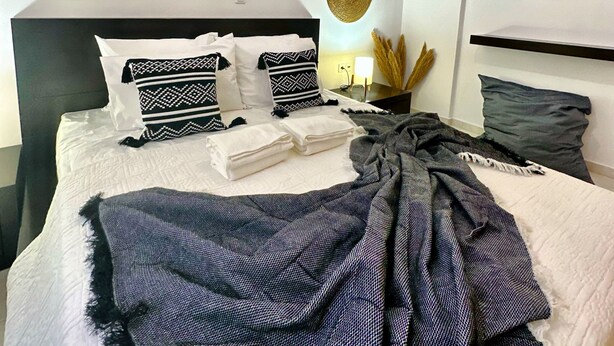 A modern bedroom with a large bed, multiple pillows, a bedside table with a lamp, and wall decor including woven circles and a mirror. - Mylos home (Ierapetra)