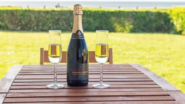 En flaske Chapel Down English Sparkling Wine og to glas på et træbord med havudsigt og grønne omgivelser i baggrunden.