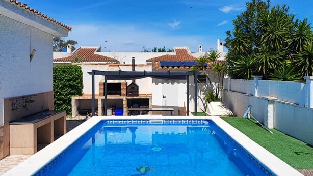 A swimming pool with a pergola and barbecue area, surrounded by a white building with a tiled roof.