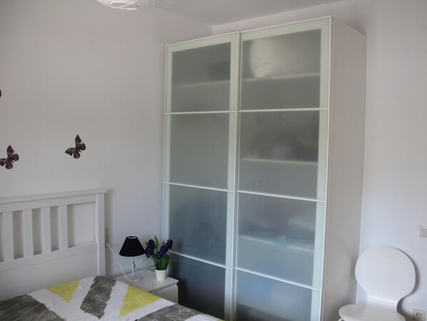 A bedroom with a bed, a white wardrobe, a bedside table with a lamp and flowers, and a white toilet.