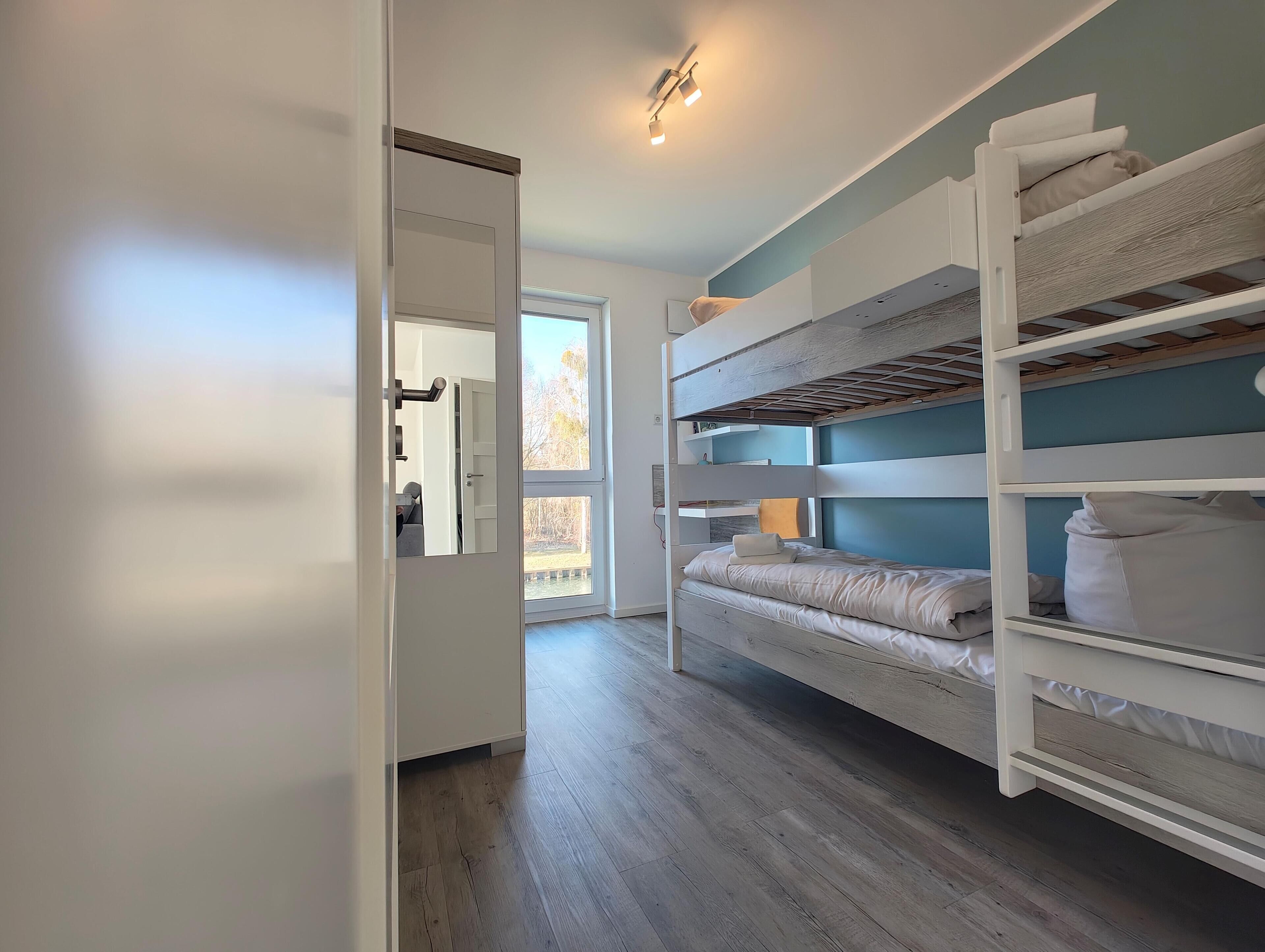 A room with a bunk bed, a door leading to another room, and a window with a view of trees.