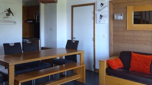 Ett kompakt vardagsrum med ett matbord i trä, svarta stolar, en soffa med röda kuddar, en köksdel och en dörr som leder till ett annat rum.