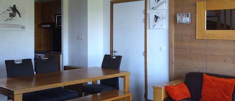 A compact living space with a wooden dining table, black chairs, a sofa with red cushions, a kitchen area, and a door leading to another room.