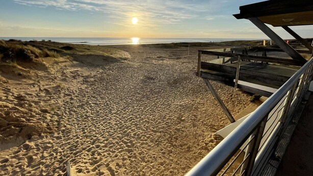 Ein Strand mit einer Holzpromenade, die zum Wasser führt, bewölkter Himmel und eine untergehende Sonne in der Ferne.