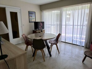 A dining area with a round table, chairs, a TV, and a large window with sheer curtains.