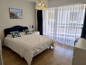 Ein Schlafzimmer mit einem Bett, einem Nachttisch, einer Lampe, einem Fenster mit transparenten Vorhängen und einem Wandbild.