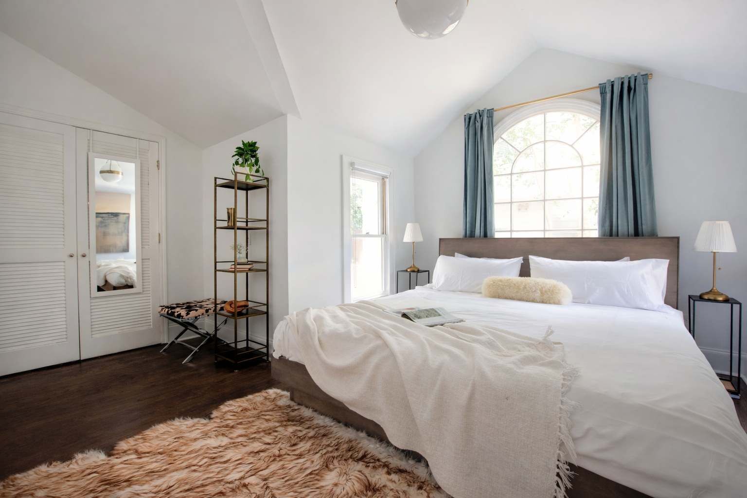 A bedroom with a large bed, a wooden floor, a white wardrobe, a window with curtains, and a plant on a shelf.