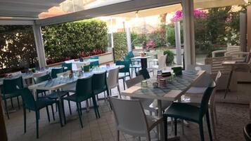 Un área de comedor al aire libre con mesas y sillas, rodeada de follaje y una sección cubierta.