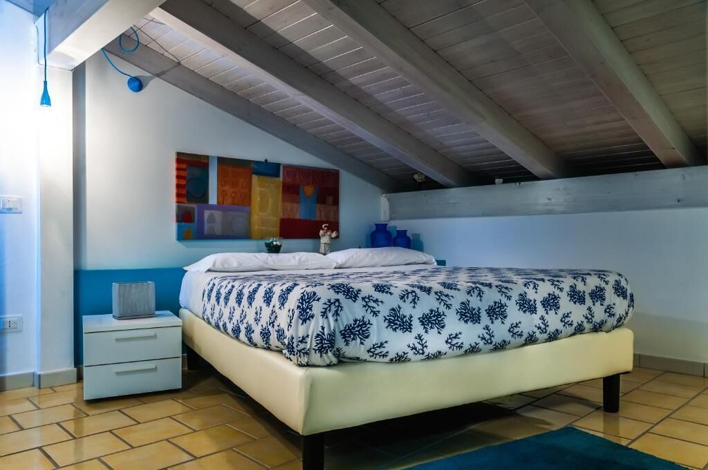 A bedroom with a bed, a nightstand, a wall with colorful artwork, and a ceiling with exposed beams.