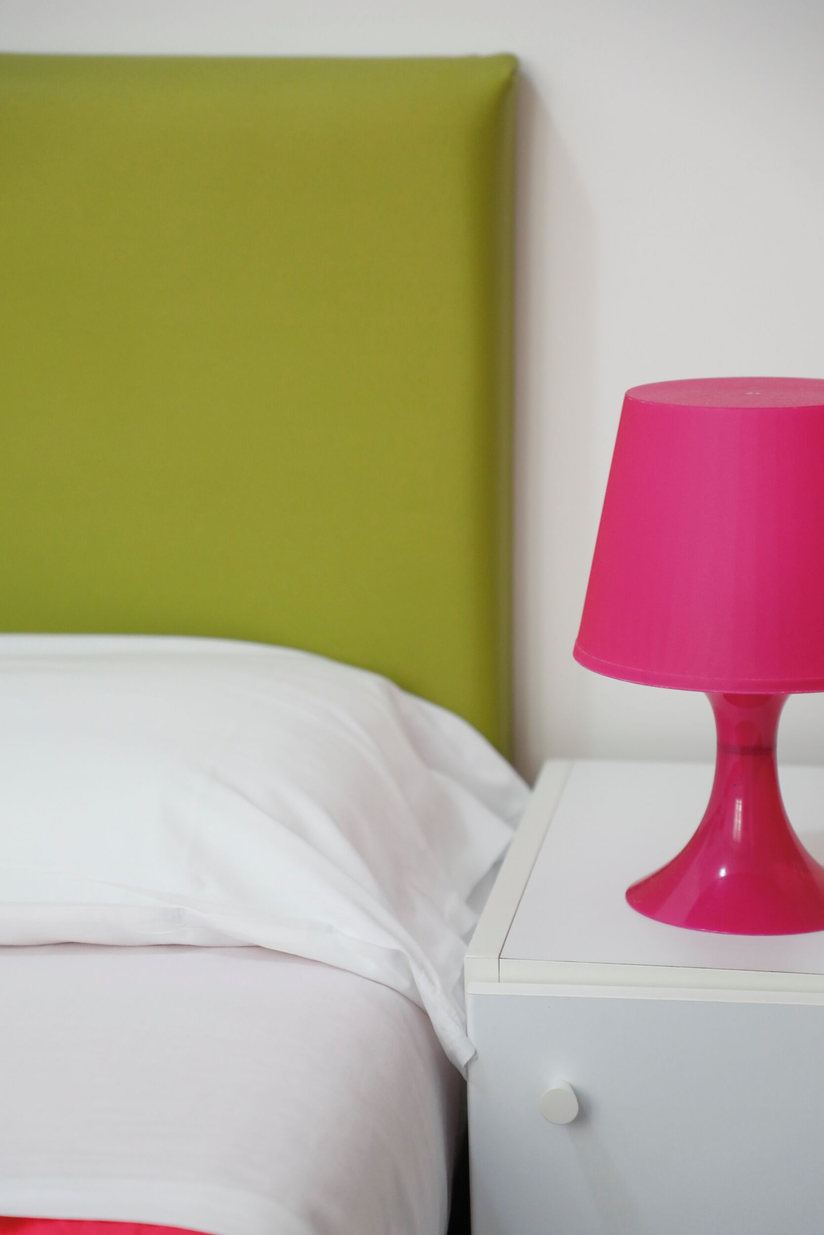 A bed with white bedding, a green headboard, and a white nightstand with a pink lamp.