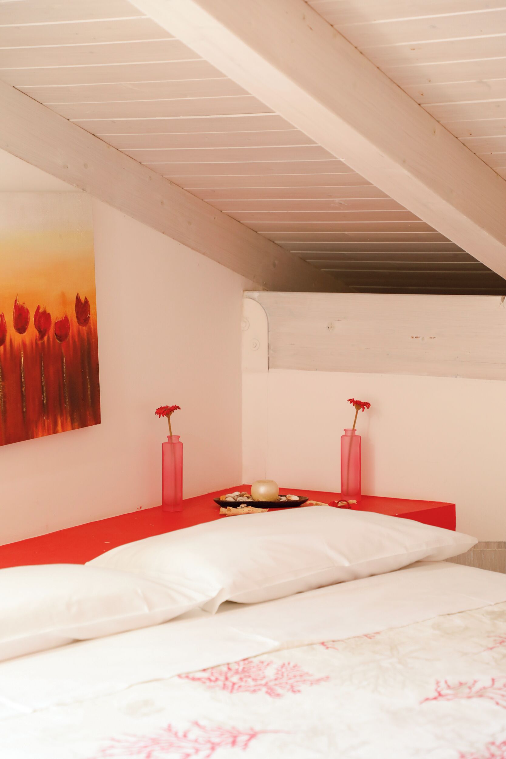 Um quarto com uma cama, uma mesa de cabeceira vermelha, dois vasos cor-de-rosa e um quadro com flores vermelhas na parede.