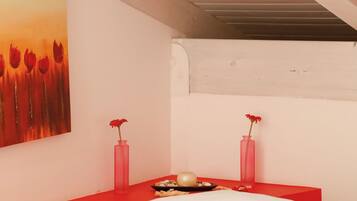 Um quarto com uma cama, uma mesa de cabeceira vermelha, dois vasos cor-de-rosa e um quadro com flores vermelhas na parede.