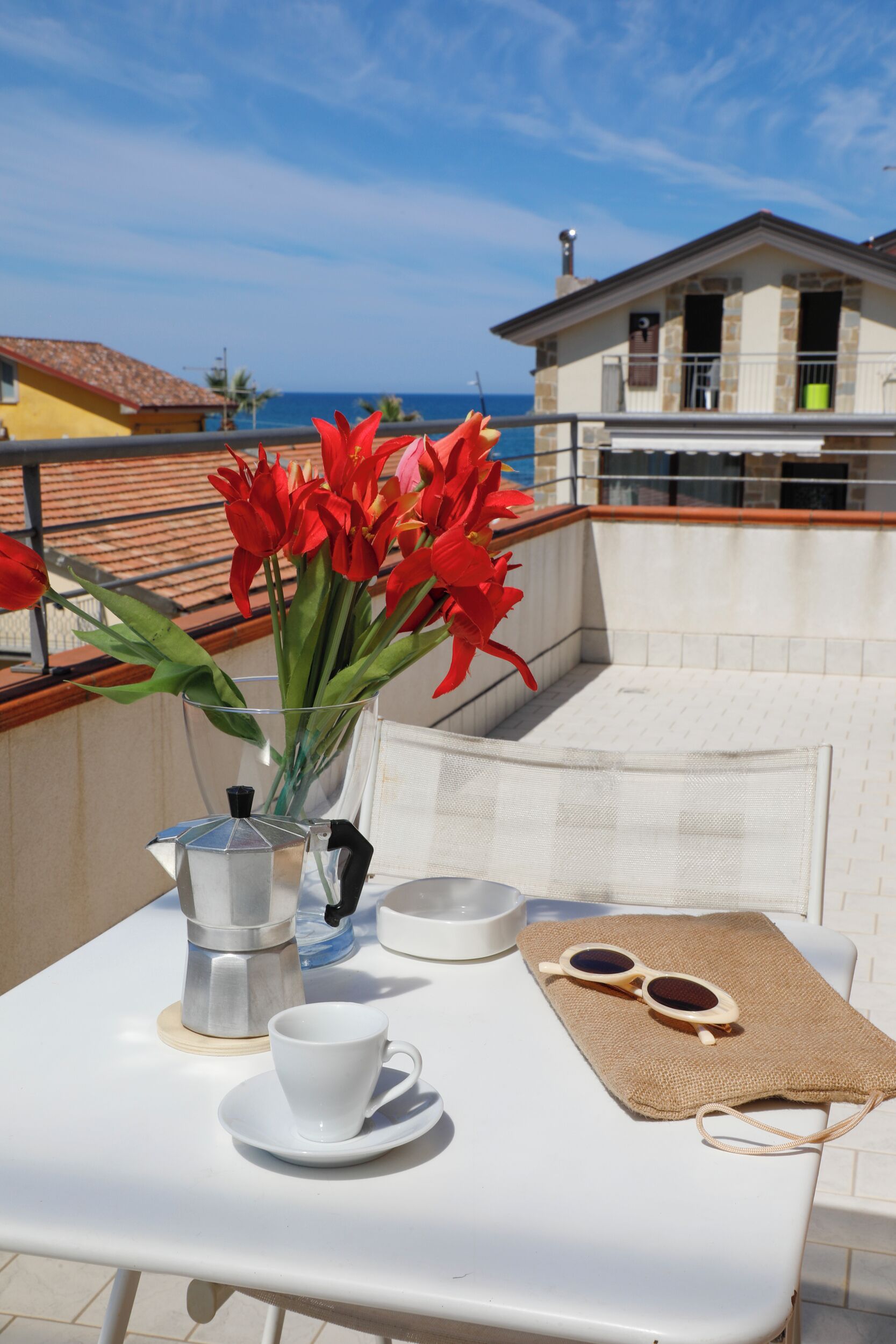 Una taula amb una cafetera, una tassa, ulleres de sol i un ram de flors vermelles en una terrassa amb vista al mar i edificis.