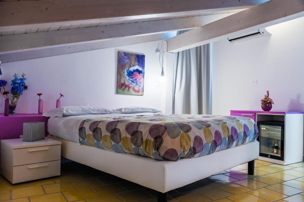 A bedroom with a bed, bedside tables, a small refrigerator, and a painting on the wall.