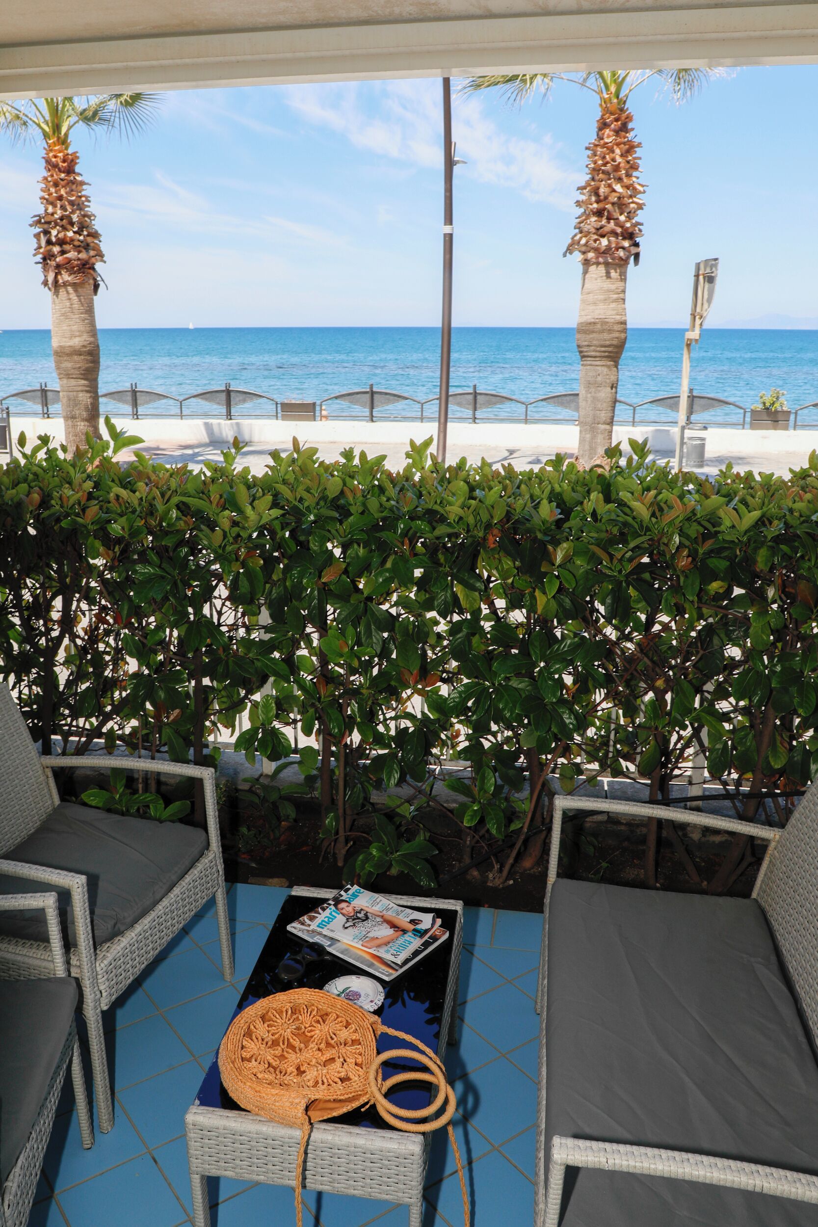 A patio with wicker furniture, a table with magazines and a woven bag, and a view of the sea.